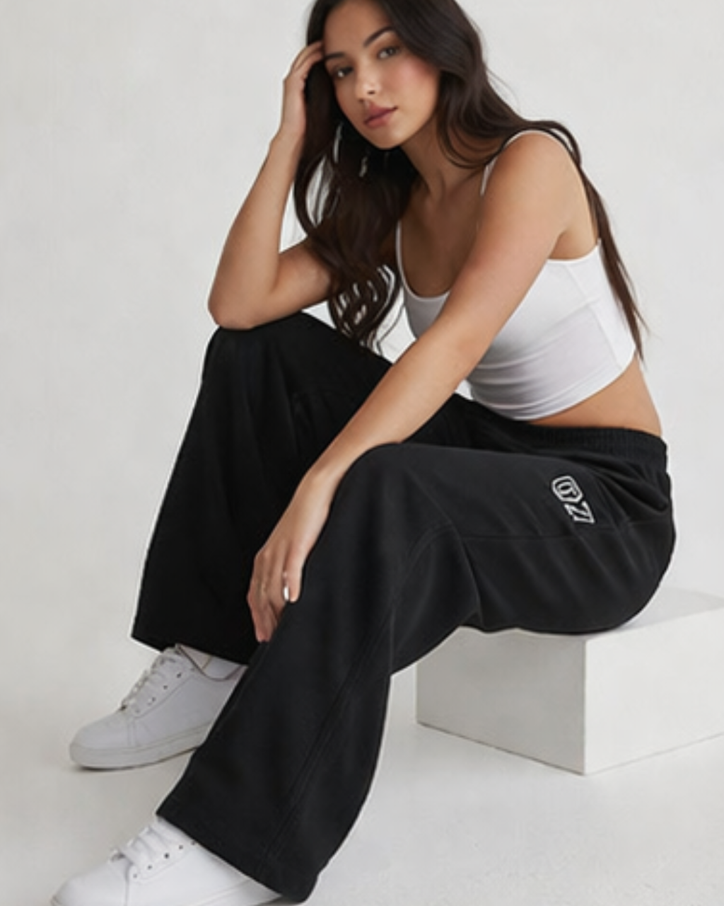Woman wearing a white tank top and black pants with a logo, sitting on a white surface.