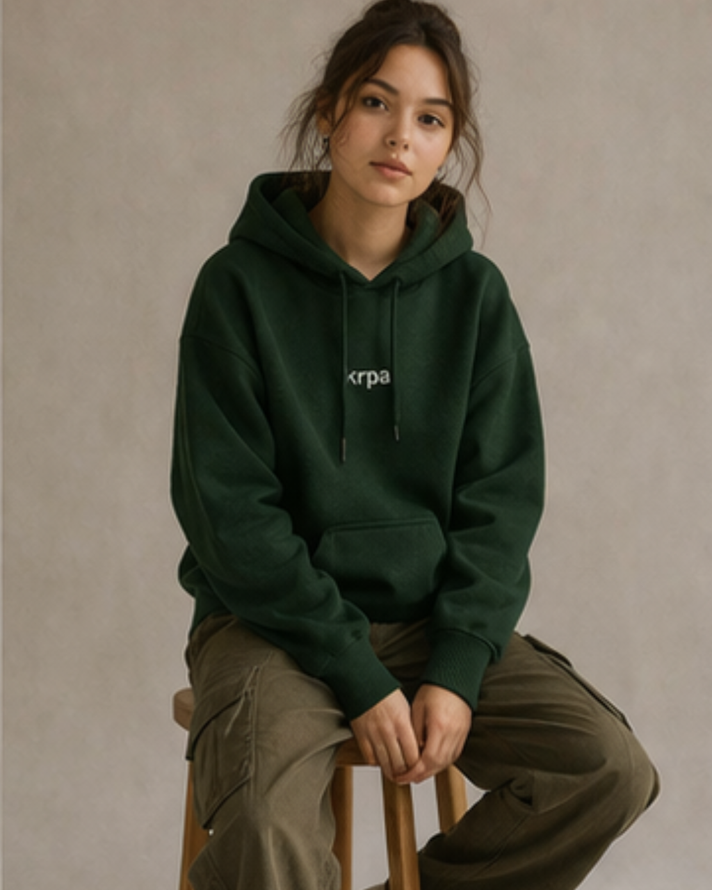 Person wearing a bottle green hoodie with krpa logo, sitting on a stool against a beige background
