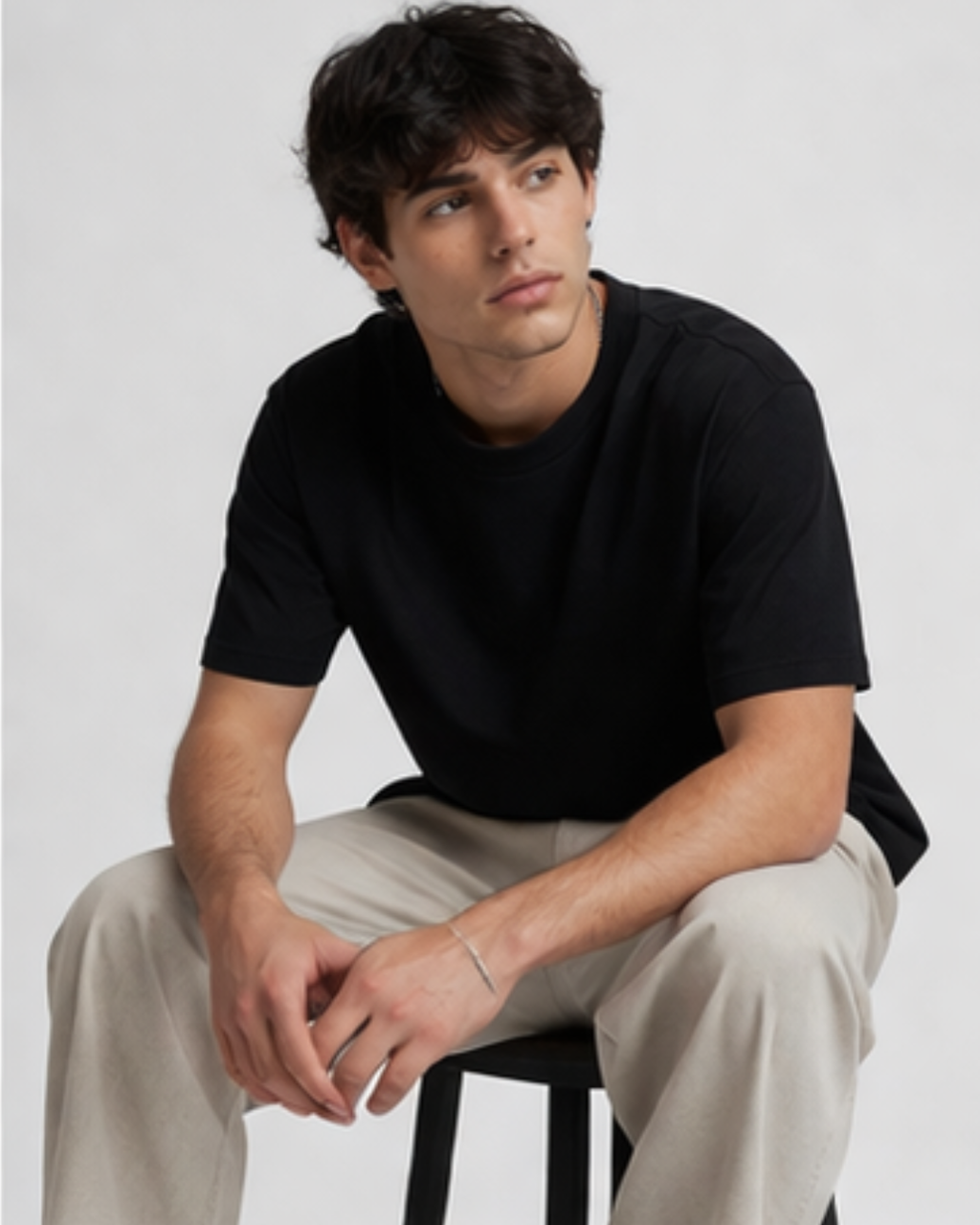 Man wearing a black t-shirt and beige pants sitting on a stool against a plain background