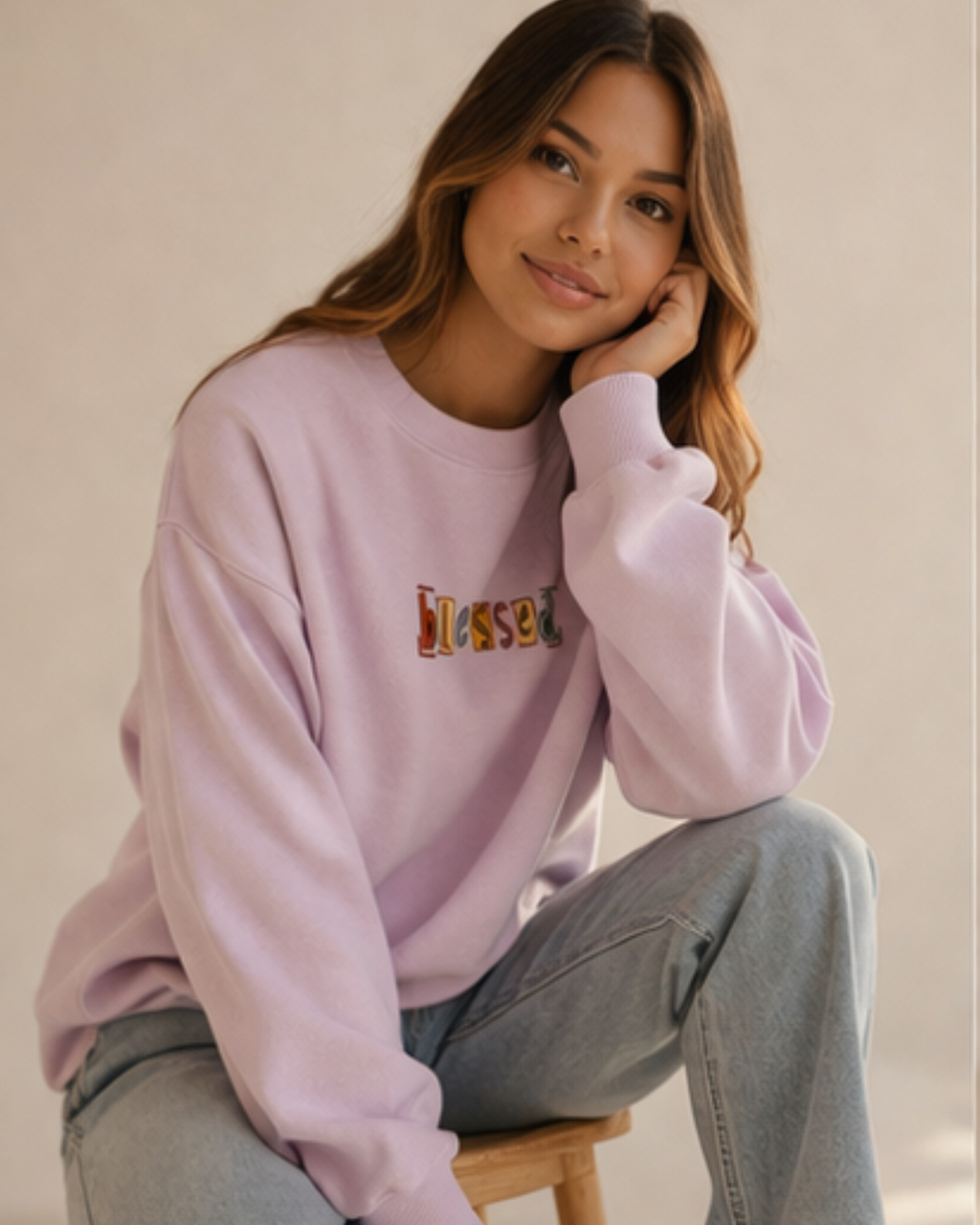 Woman wearing a baby pink sweatshirt with colorful text sitting on a wooden stool against a beige wall.