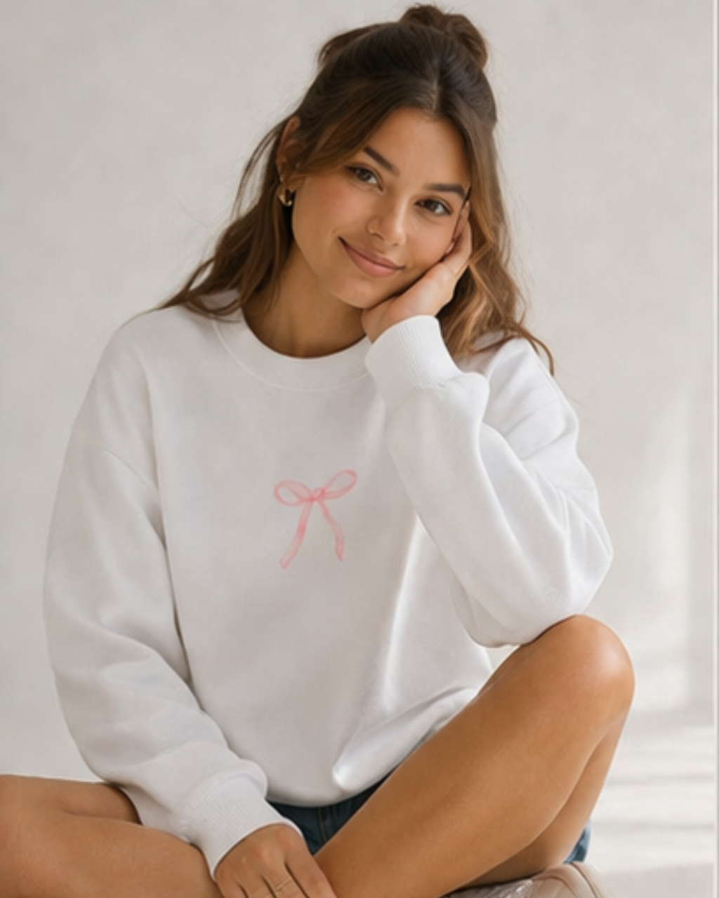 Woman wearing a white sweatshirt with a pink bow design, sitting against a light background.