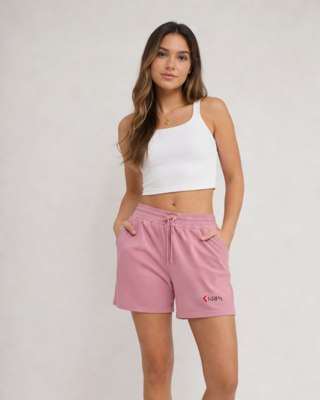 Woman wearing a white crop top and pink shorts with KRPA logo on a plain background