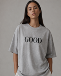 Person wearing a grey oversized t-shirt with 'GOOD' printed on it against a plain background