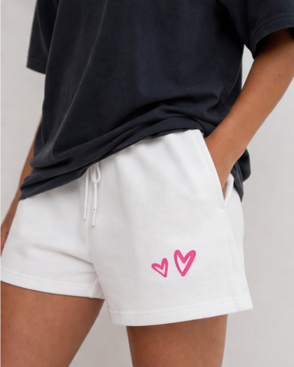 Person wearing white shorts with pink heart designs and a black top on a light gray background