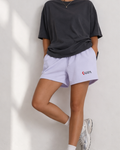 Person wearing a black oversized t-shirt and light purple shorts with a brand logo against a white wall.
