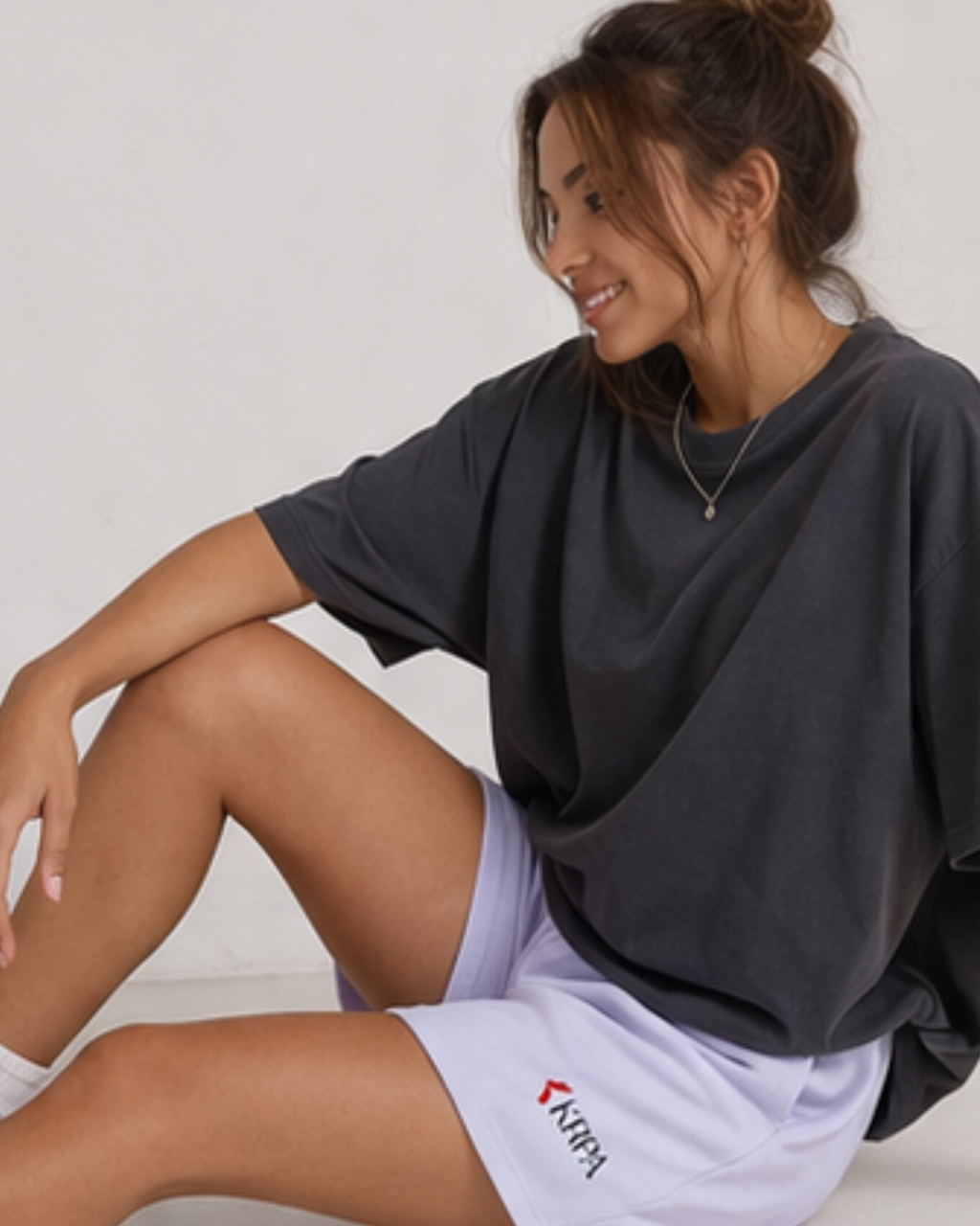 Woman wearing a dark gray oversized shirt and white shorts with a brand logo, sitting on a light-colored floor.