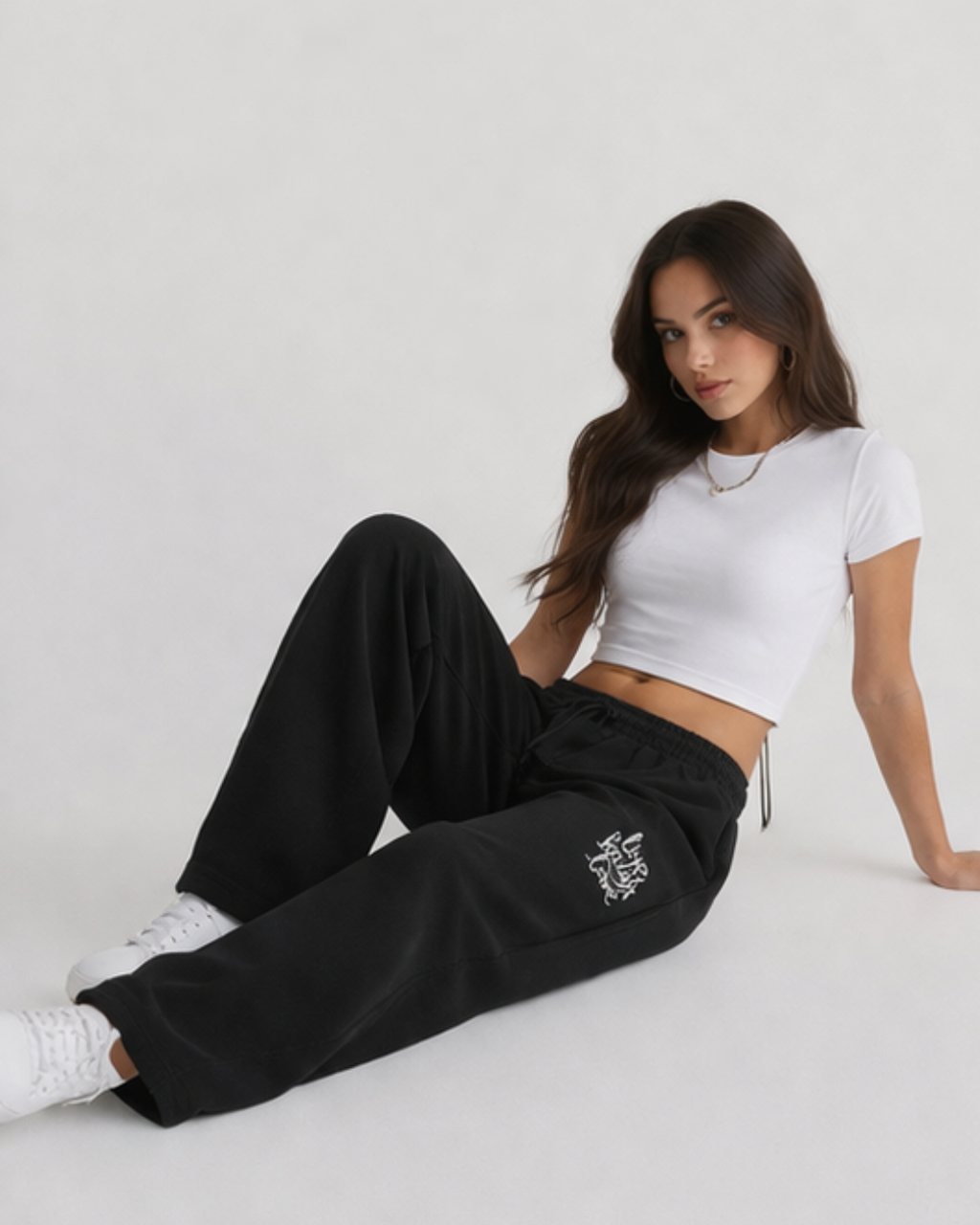 Woman wearing a white crop top and black pants with a logo, sitting on a light gray background.