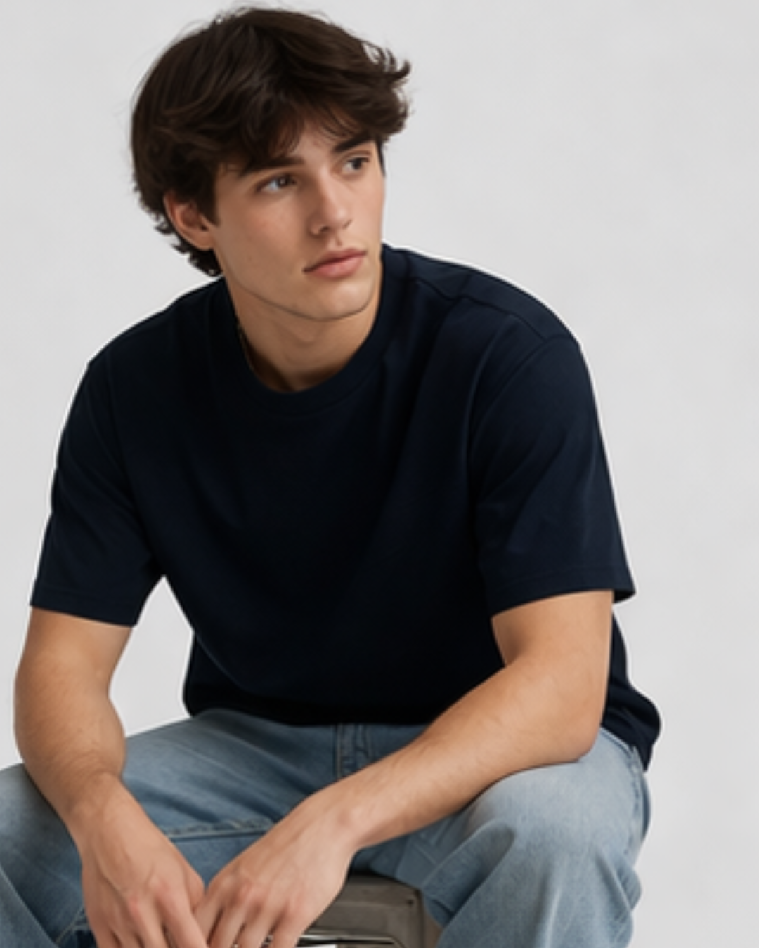 Man wearing a navy-blue t-shirt and light blue jeans sitting on a chair against a plain background
