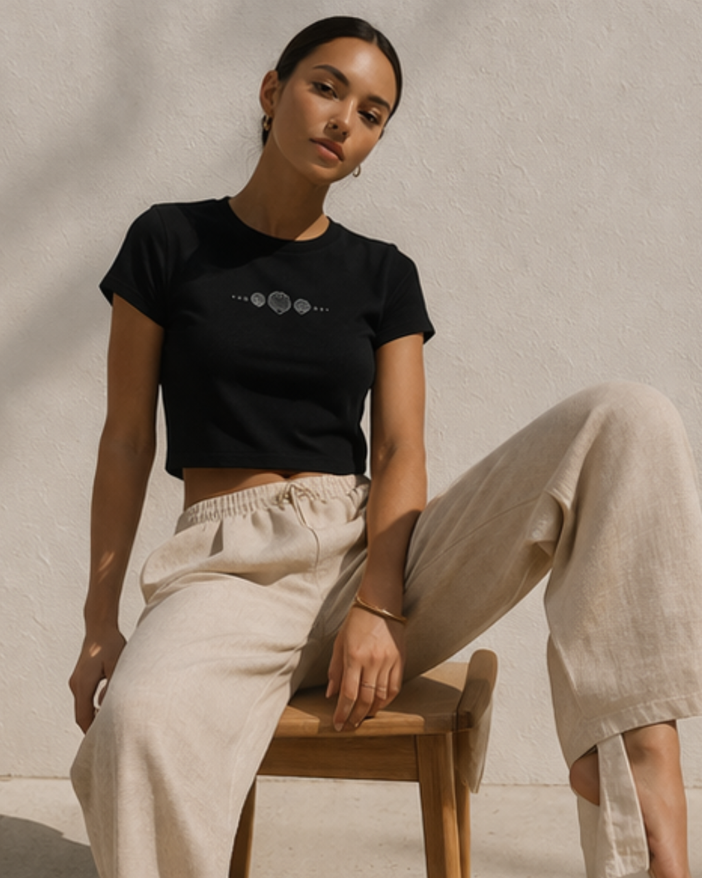 Woman wearing a black cropped t-shirt and beige pants sitting on a wooden stool against a neutral background