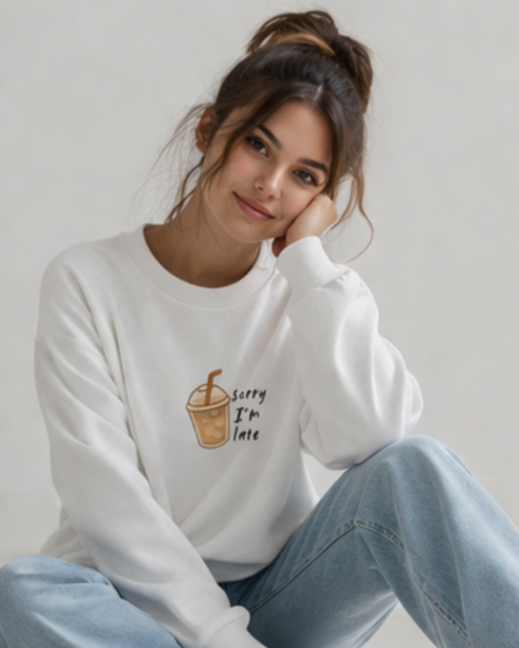 a person sitting on the floor wearing white sweatshirt which says 'sorry I'm late' paired with blue jeans on a plain light background.