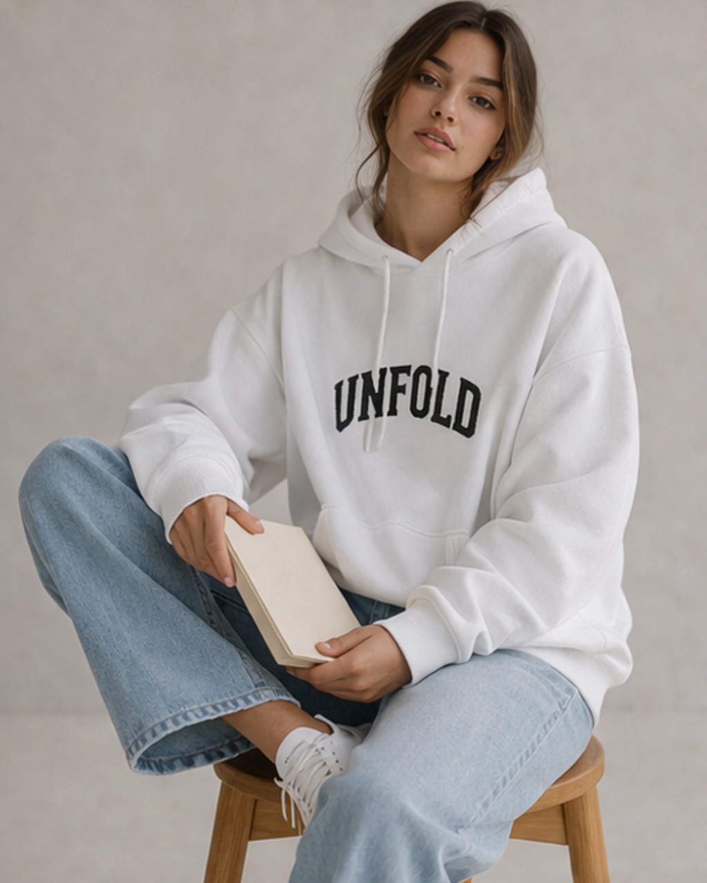 Person wearing a white hoodie with 'UNFOLD' text, sitting on a stool holding a book.